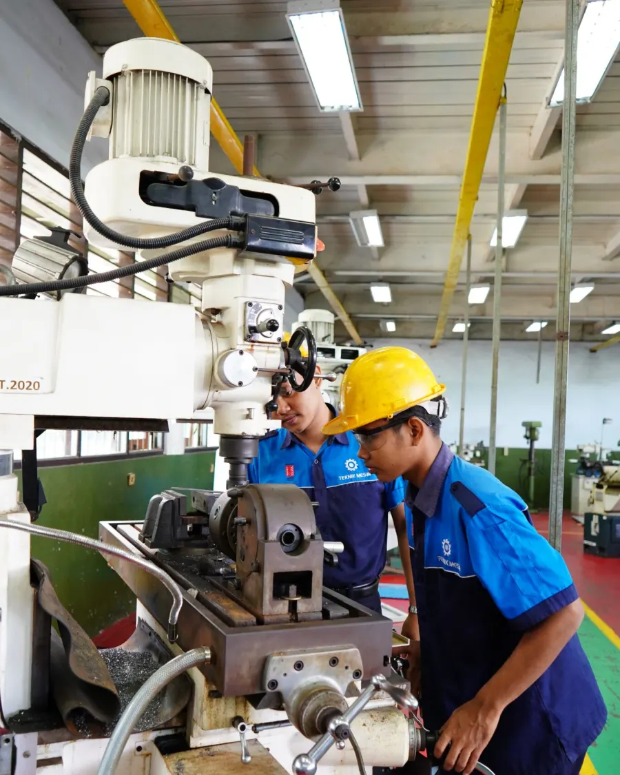 Dua siswa TPM berhelm dan berkacamata pelindung menyetel benda kerja di mesin frais bengkel TPM